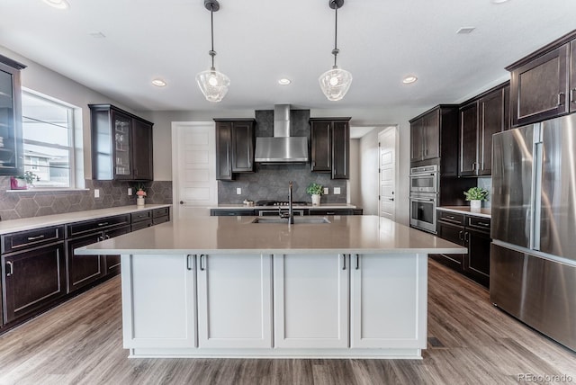 kitchen with an island with sink, a sink, appliances with stainless steel finishes, wall chimney exhaust hood, and glass insert cabinets