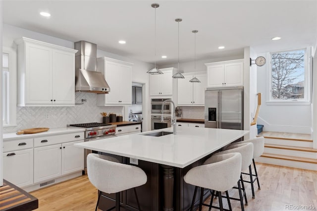 kitchen with a center island with sink, white cabinets, wall chimney range hood, hanging light fixtures, and appliances with stainless steel finishes