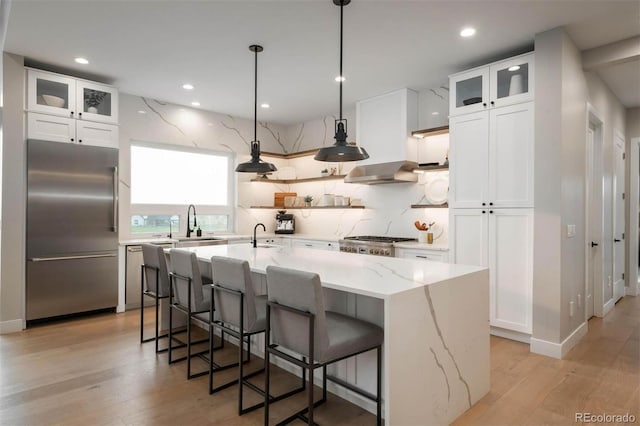 kitchen with light stone countertops, stainless steel appliances, a center island with sink, white cabinetry, and range hood