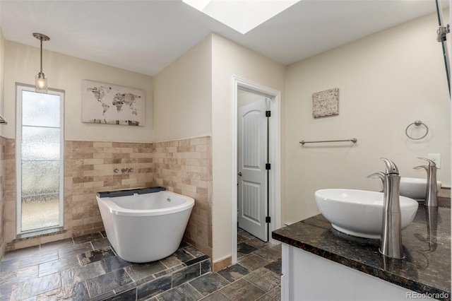 bathroom with vanity, stone tile floors, a skylight, a freestanding tub, and tile walls