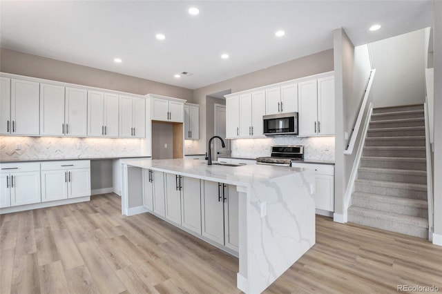 kitchen with sink, an island with sink, white cabinetry, light wood-type flooring, and appliances with stainless steel finishes