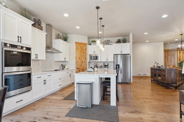 kitchen with appliances with stainless steel finishes, an island with sink, white cabinets, hanging light fixtures, and wall chimney range hood