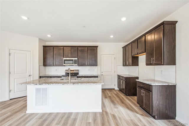 kitchen with appliances with stainless steel finishes, an island with sink, light stone counters, light hardwood / wood-style floors, and dark brown cabinets