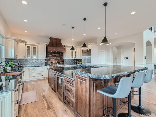 kitchen with a large island, decorative light fixtures, custom range hood, backsplash, and dark stone countertops