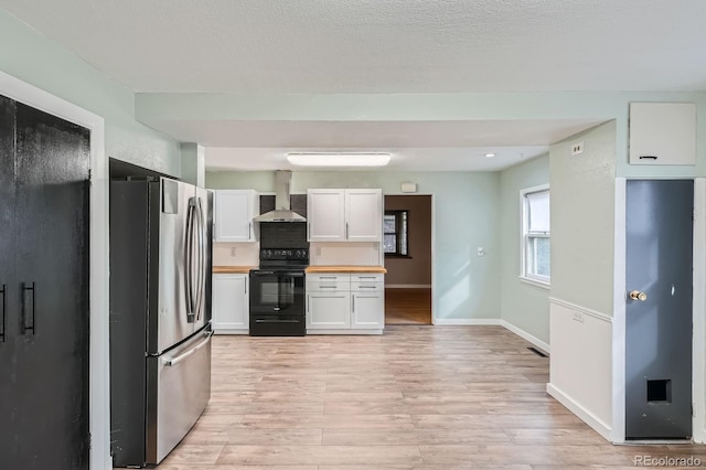 kitchen with black electric range oven, light wood-style flooring, freestanding refrigerator, white cabinetry, and wall chimney range hood