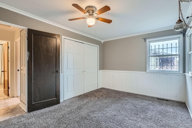 unfurnished bedroom featuring carpet floors, a wainscoted wall, crown molding, visible vents, and a ceiling fan