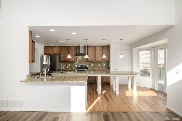 kitchen featuring wall chimney range hood, a breakfast bar area, appliances with stainless steel finishes, hanging light fixtures, and backsplash