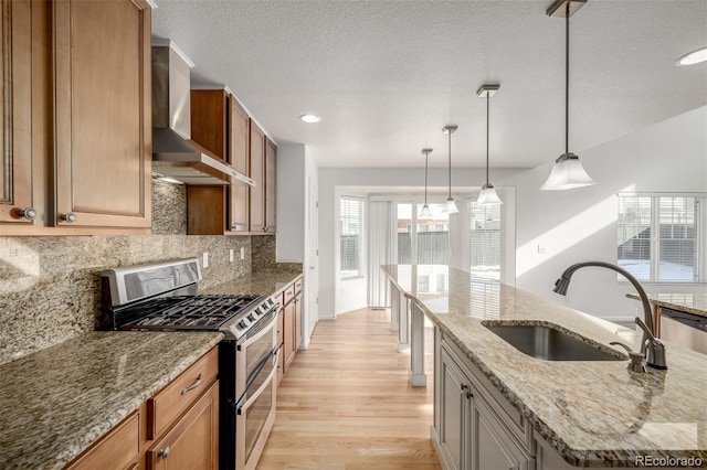 kitchen with wall chimney range hood, sink, appliances with stainless steel finishes, light stone countertops, and an island with sink