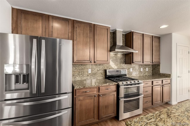 kitchen featuring wall chimney exhaust hood, light stone counters, stainless steel appliances, light hardwood / wood-style floors, and decorative backsplash