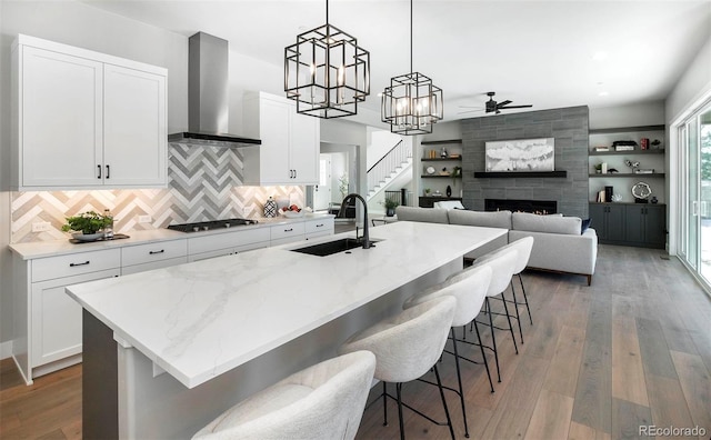 kitchen featuring stainless steel gas stovetop, white cabinets, hanging light fixtures, a center island with sink, and wall chimney exhaust hood