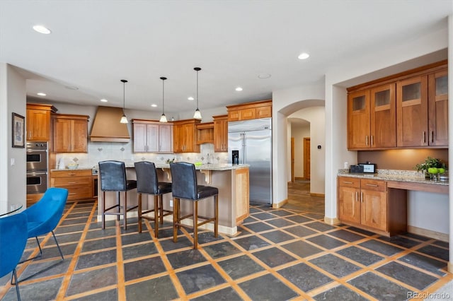 kitchen with appliances with stainless steel finishes, custom exhaust hood, brown cabinetry, recessed lighting, and arched walkways