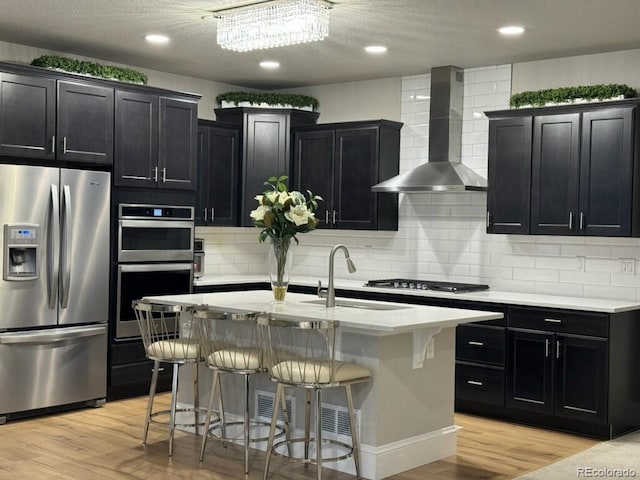 kitchen with a kitchen breakfast bar, appliances with stainless steel finishes, wall chimney exhaust hood, and a center island with sink