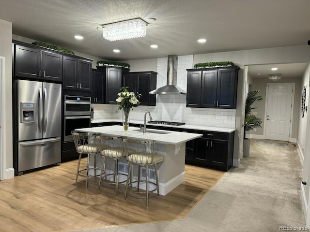 kitchen with a center island with sink, light wood-type flooring, appliances with stainless steel finishes, a kitchen bar, and wall chimney exhaust hood