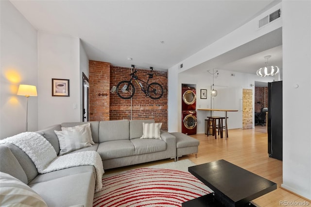 living area featuring visible vents, brick wall, light wood finished floors, an inviting chandelier, and stacked washer / drying machine