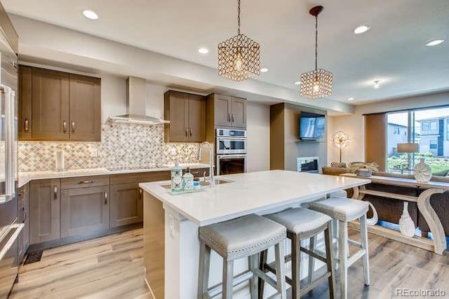 kitchen with sink, wall chimney exhaust hood, double oven, light hardwood / wood-style floors, and a breakfast bar area