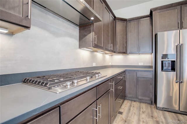 kitchen with wall chimney range hood, appliances with stainless steel finishes, dark brown cabinets, and light hardwood / wood-style floors