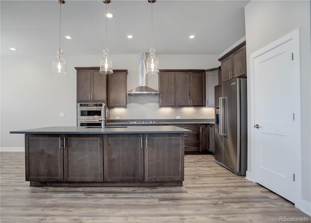 kitchen featuring light wood-type flooring, wall chimney range hood, appliances with stainless steel finishes, dark brown cabinets, and hanging light fixtures