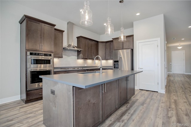 kitchen featuring wall chimney exhaust hood, sink, a kitchen island with sink, light wood-type flooring, and appliances with stainless steel finishes