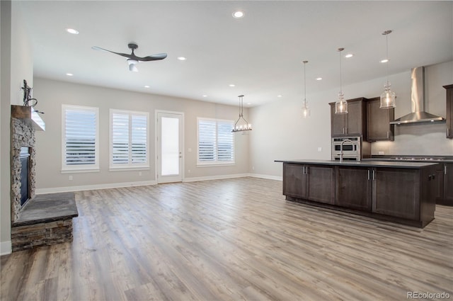 kitchen with a stone fireplace, wall chimney range hood, light wood-type flooring, dark brown cabinets, and hanging light fixtures