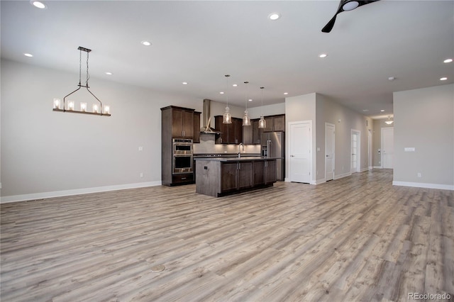 kitchen featuring stainless steel appliances, wall chimney range hood, light wood-type flooring, pendant lighting, and an island with sink