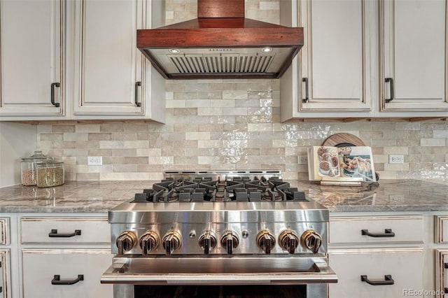 kitchen featuring light stone counters, range hood, decorative backsplash, white cabinetry, and high end stove