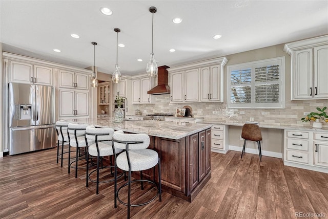 kitchen featuring dark wood-style flooring, built in desk, stainless steel fridge with ice dispenser, wall chimney range hood, and a kitchen bar