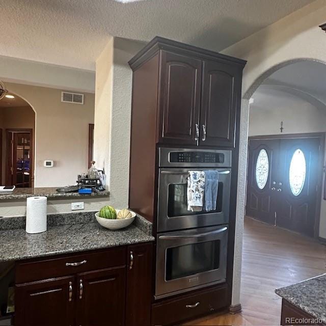 13291 N Harback Rd, Bennett, CO 80102 - kitchen with double oven, arched walkways, dark stone counters, light wood finished floors, and a textured ceiling