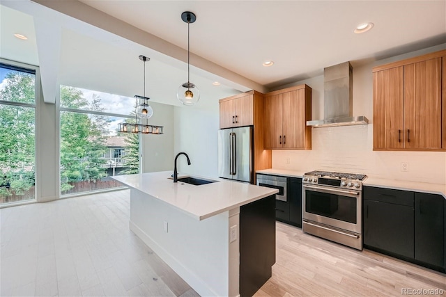 kitchen featuring a kitchen island with sink, sink, wall chimney exhaust hood, appliances with stainless steel finishes, and decorative light fixtures