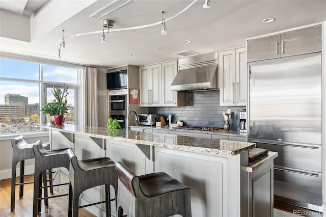 kitchen with decorative backsplash, wall chimney exhaust hood, light wood-style flooring, light stone countertops, and stainless steel appliances