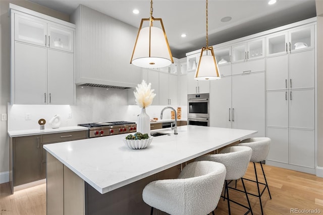 kitchen featuring a center island with sink, white cabinets, hanging light fixtures, and double oven