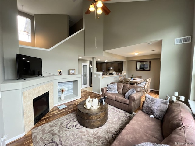 living room featuring ceiling fan, a towering ceiling, hardwood / wood-style floors, and a tile fireplace