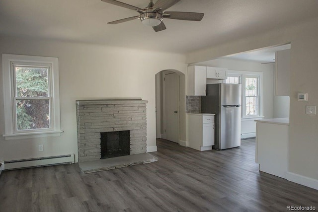 unfurnished living room with dark wood-style floors, baseboard heating, a fireplace, and a ceiling fan