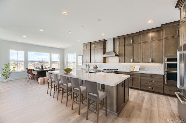 kitchen featuring stainless steel appliances, wall chimney exhaust hood, an island with sink, a kitchen breakfast bar, and light hardwood / wood-style flooring