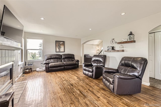 living area with arched walkways, wood-type flooring, a tiled fireplace, recessed lighting, and baseboards