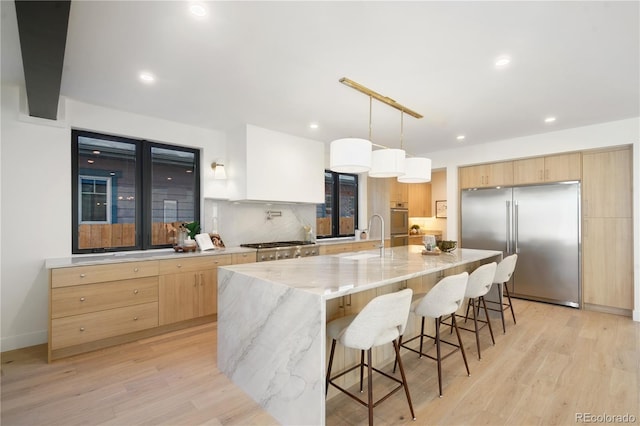 kitchen featuring a large island with sink, built in fridge, sink, decorative light fixtures, and light hardwood / wood-style floors