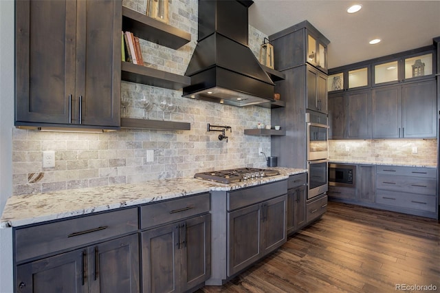 kitchen featuring light stone counters, dark wood-style floors, glass insert cabinets, appliances with stainless steel finishes, and premium range hood