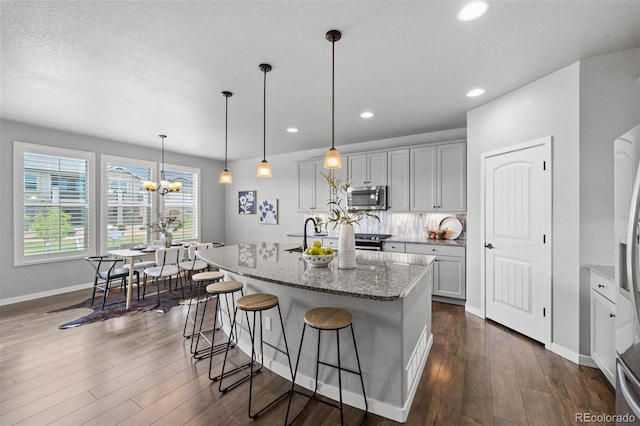 kitchen featuring dark wood-type flooring, an inviting chandelier, a center island with sink, hanging light fixtures, and light stone counters