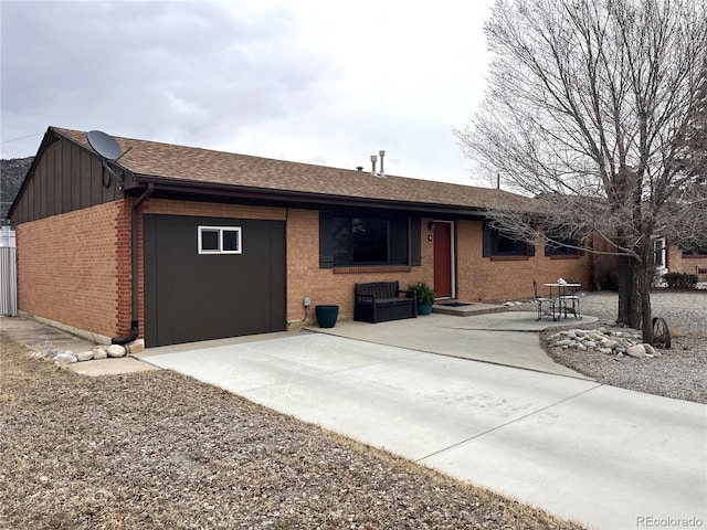 single story home with a garage, brick siding, a shingled roof, board and batten siding, and a patio area