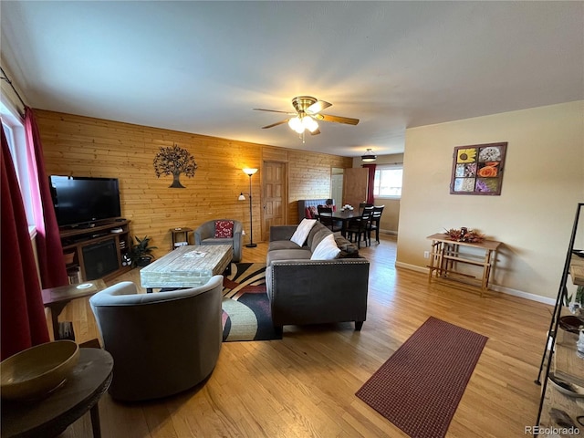 living area featuring light wood-style floors, wood walls, baseboards, and a ceiling fan