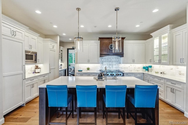 kitchen featuring stainless steel microwave, a center island with sink, decorative backsplash, and light hardwood / wood-style floors