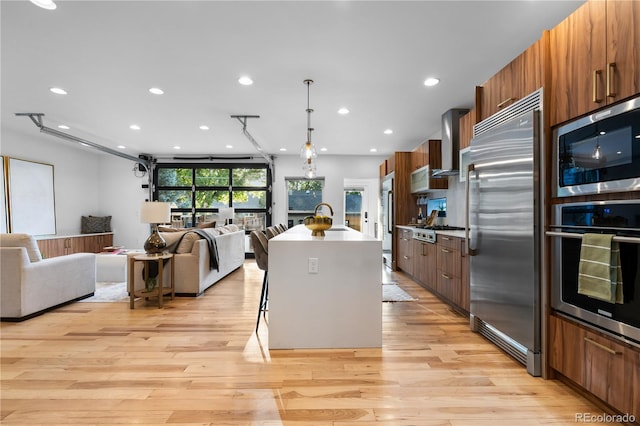 kitchen with an island with sink, a breakfast bar area, built in appliances, light hardwood / wood-style flooring, and wall chimney range hood