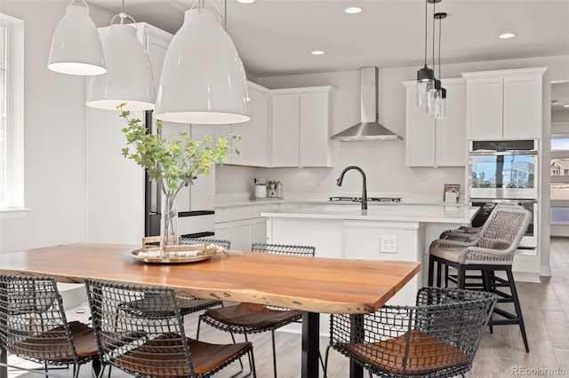 kitchen with double oven, an island with sink, light wood-type flooring, light countertops, and wall chimney exhaust hood