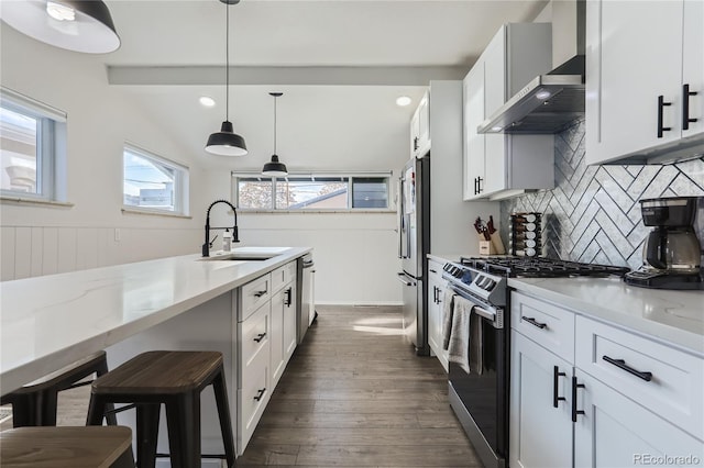 kitchen with wall chimney range hood, dark wood finished floors, pendant lighting, stainless steel appliances, and a sink