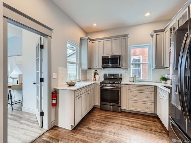 kitchen featuring light stone counters, gray cabinetry, stainless steel appliances, wood finished floors, and decorative backsplash