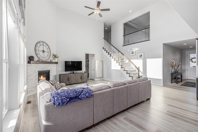 living area with a ceiling fan, a towering ceiling, light wood-style flooring, stairway, and a stone fireplace