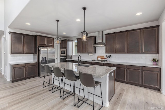 kitchen with wall chimney exhaust hood, light wood-style flooring, a breakfast bar, stainless steel appliances, and a sink