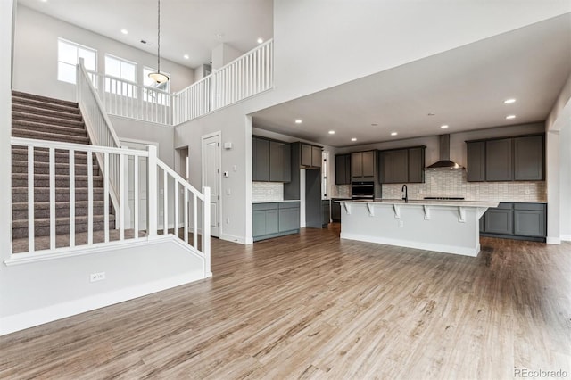 kitchen featuring stainless steel oven, wall chimney exhaust hood, backsplash, an island with sink, and light hardwood / wood-style floors