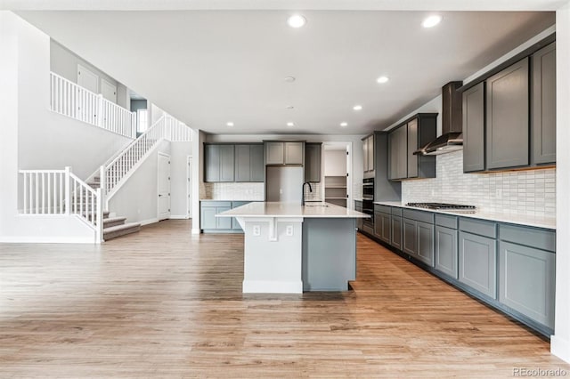 kitchen with an island with sink, light hardwood / wood-style floors, wall chimney range hood, and stainless steel gas cooktop