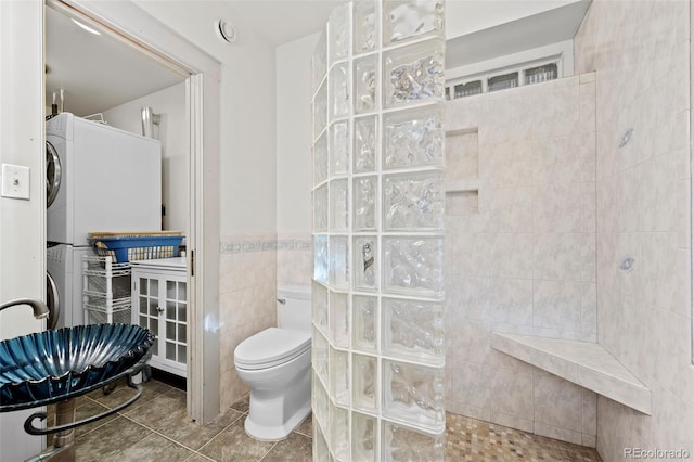 bathroom featuring tile patterned floors, tiled shower, tile walls, stacked washer and dryer, and toilet