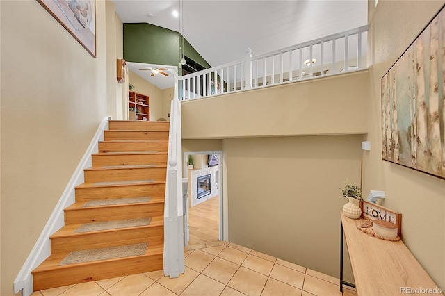 staircase with tile patterned flooring, a glass covered fireplace, ceiling fan, and high vaulted ceiling